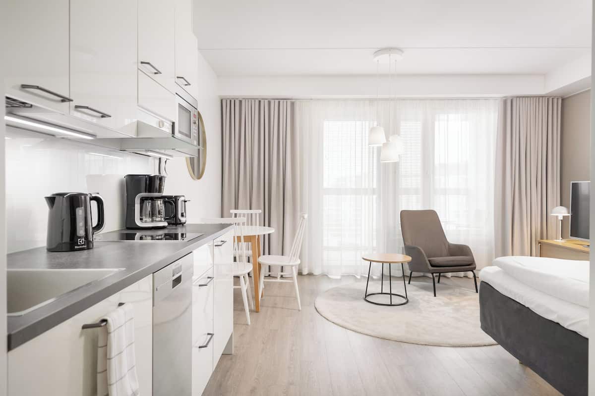 A modern hotel room with a kitchenette, a dining area, a large bed, and a view of the outdoors through sheer curtains.