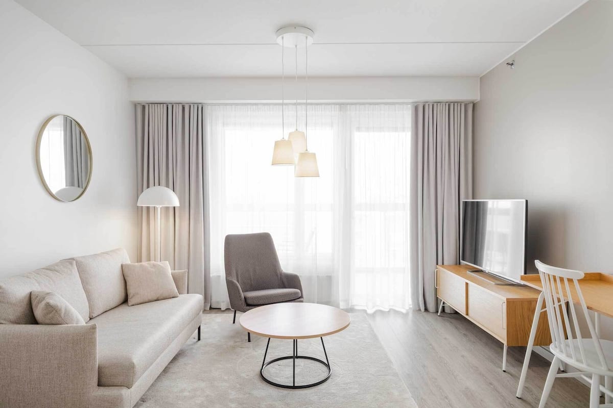 A modern hotel room with a sofa, armchair, round table, TV, and a large window with curtains.