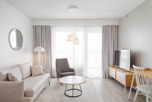 Habitación de hotel moderna con sofá, sillón, mesa redonda, televisor y un ventanal con cortinas.