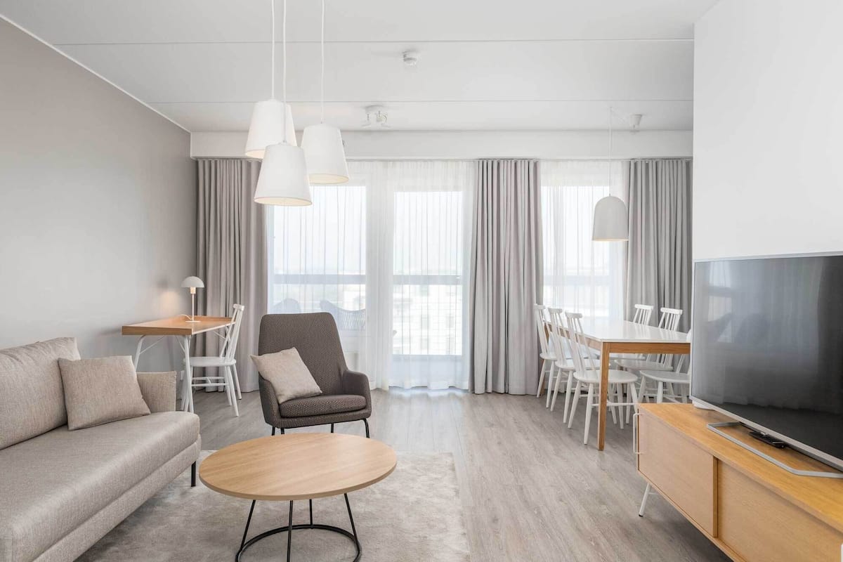 A modern hotel room with a sofa, round table, dining set, and a TV.
