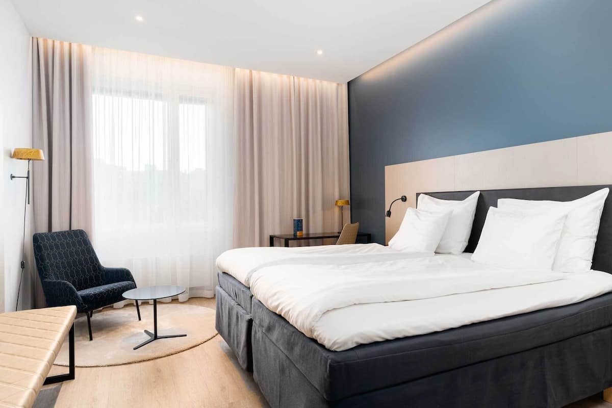 A modern hotel room with a large bed, a desk, a chair, and a sofa.