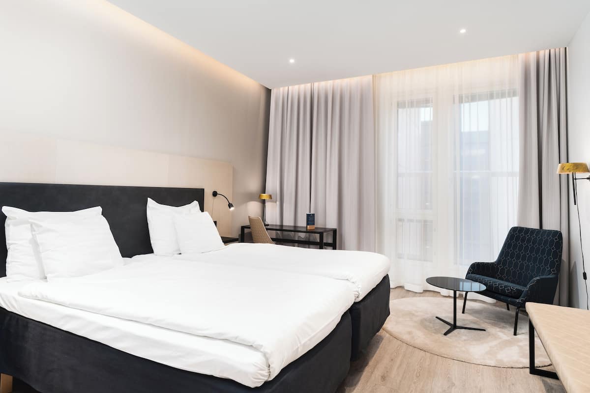 A modern hotel room with a large bed, a desk, a chair, and a small table.