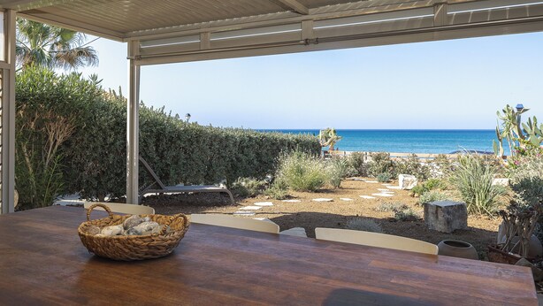Een terras in de open lucht met een houten tafel en een rieten mand, met uitzicht op een strand met helderblauw water en een rotsachtig landschap.