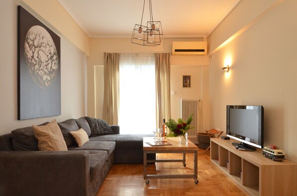 A living room with a sofa, a coffee table with a vase, a television on a wooden stand, and an air conditioning unit on the wall.
