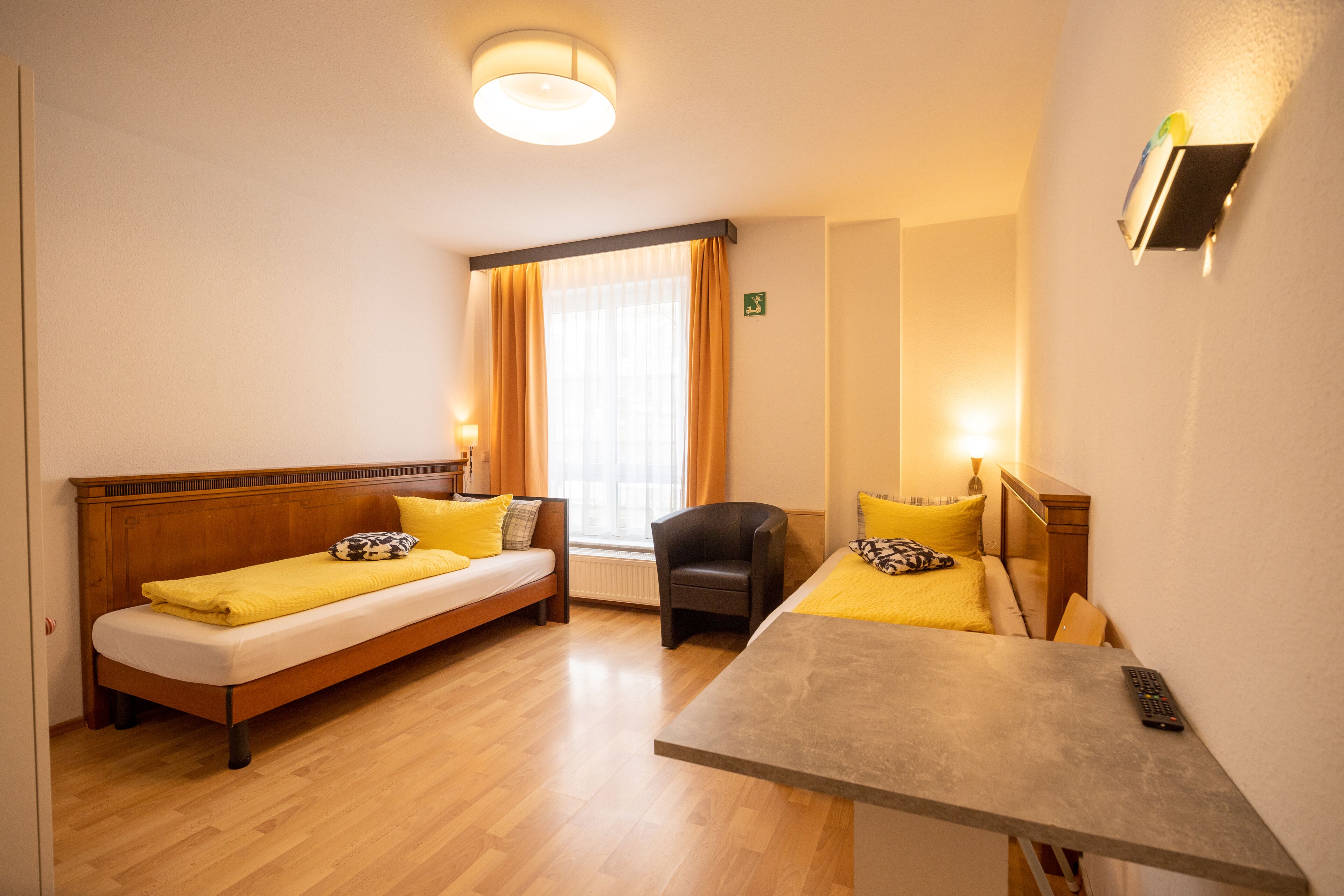 Hotellihuone, jossa on kaksi erillistä sänkyä, pieni pöytä, tuoli, ikkuna, jossa on verhot, ja seinään kiinnitetty valaisin.