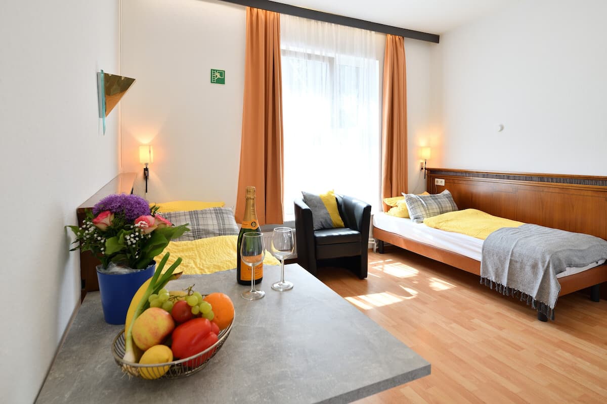 A hotel room with a bed, a chair, a table with fruit and a bottle, and a window with curtains.