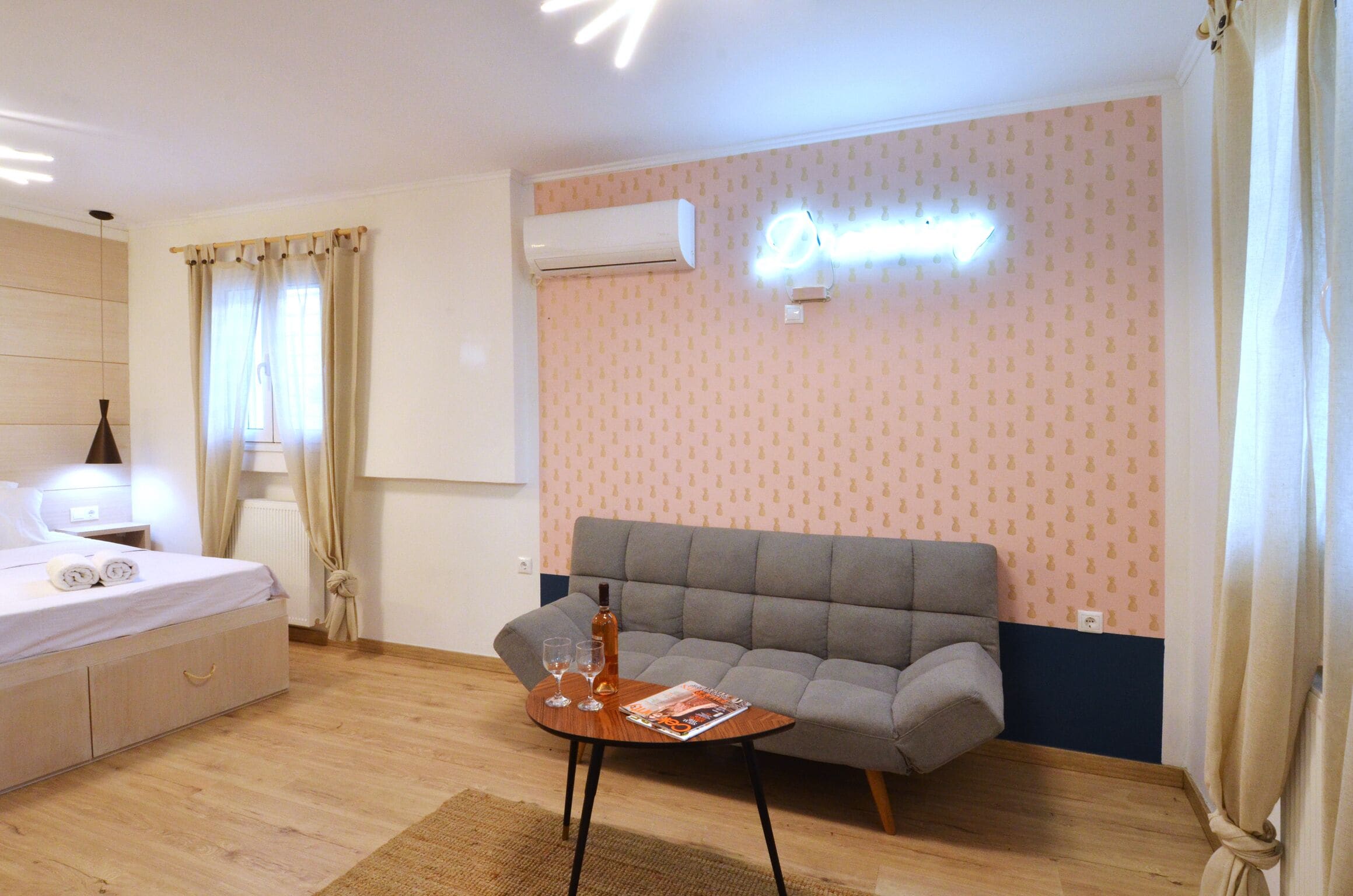 Habitación de hotel moderna con cama, un sofá gris, una mesita con una revista y un equipo de aire acondicionado en la pared.