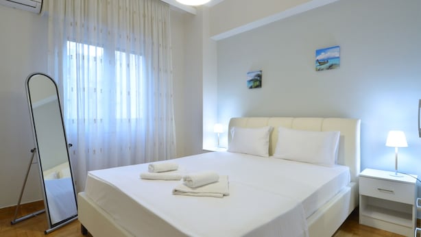 A hotel room with a large bed, a bedside table with a lamp, a mirror, and a window with curtains. - Niovis a nice and cozy apartment (Athens)