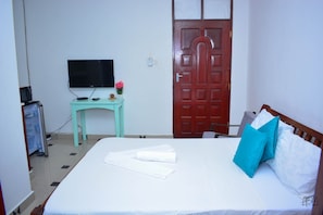 A bedroom with a bed, a television on a stand, a desk with a computer, and a door. - Nyali Studio Apartment (Mombasa)