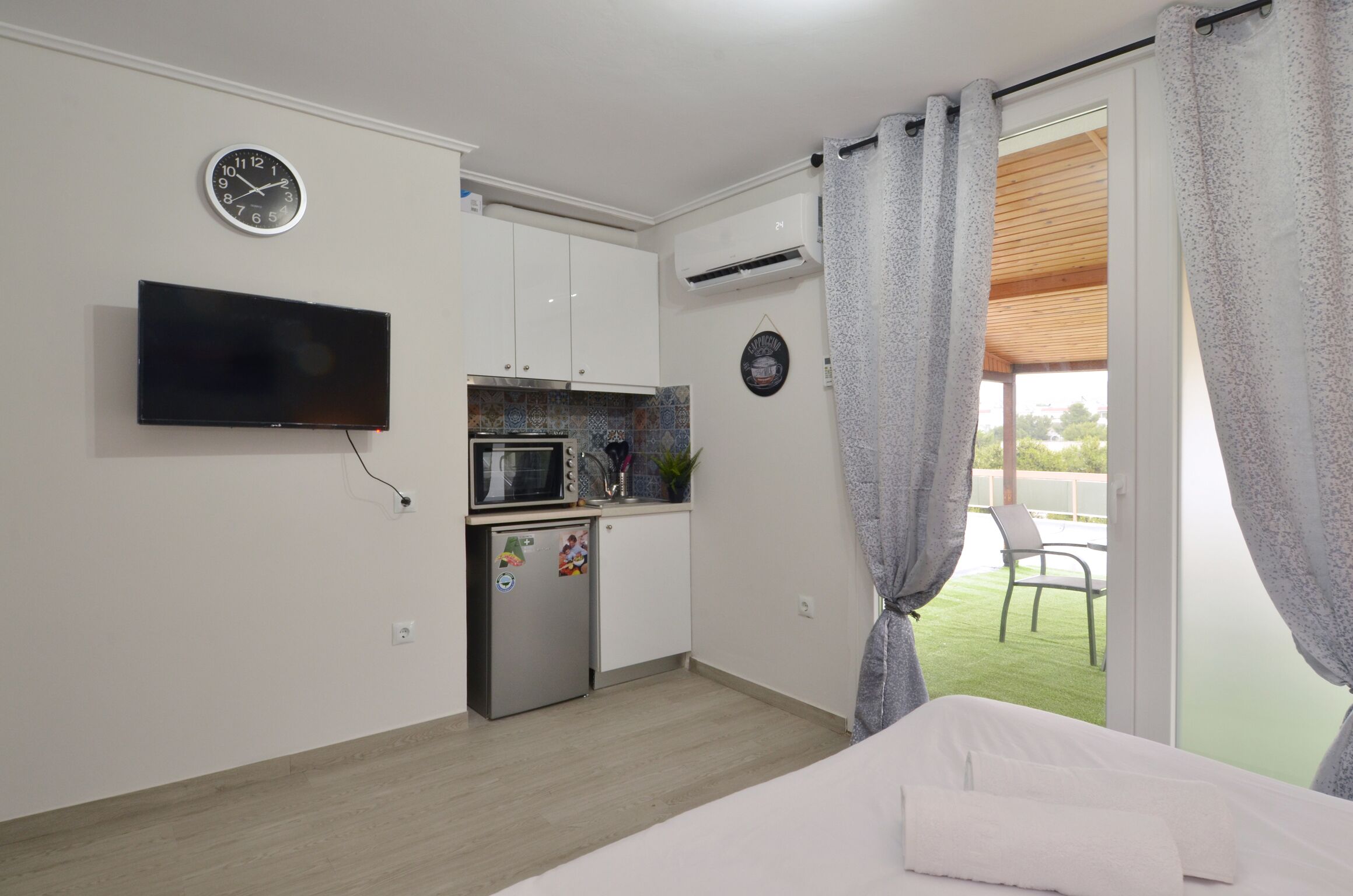 A bedroom with a bed, a TV, a kitchenette, and a balcony with curtains.