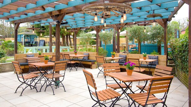 An outdoor patio with wooden tables and benches, a rustic ceiling with exposed beams, and hanging light fixtures. - The Curious Pig in the Parlour  (Crawley)