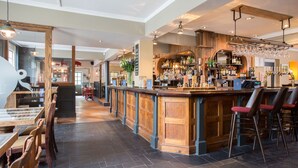 A bar area with wooden bar stools, a counter with bottles, and a seating area with tables and chairs. - The Curious Pig in the Parlour  (Crawley)