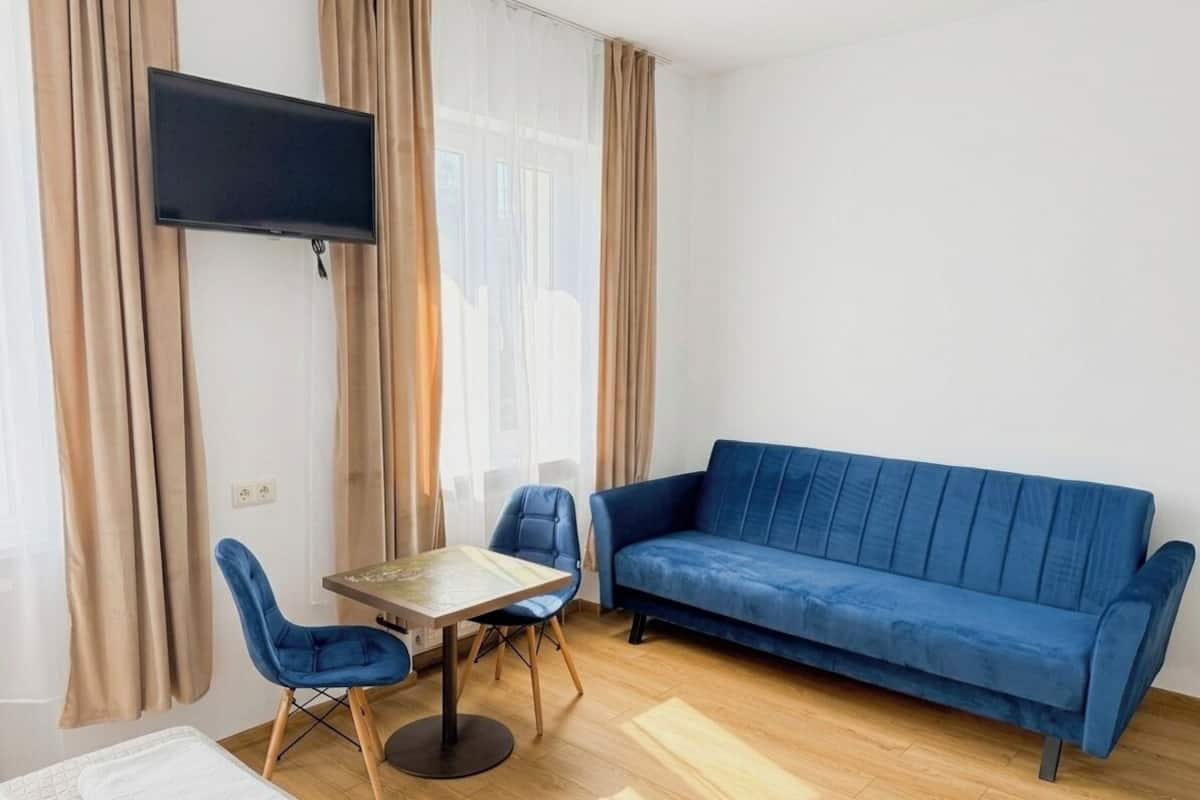 Une pièce avec un canapé bleu, une petite table entourée de chaises, un téléviseur fixé au mur et un lit recouvert d’une couverture blanche.