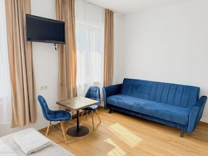 Une pièce avec un canapé bleu, une petite table entourée de chaises, un téléviseur fixé au mur et un lit recouvert d’une couverture blanche.