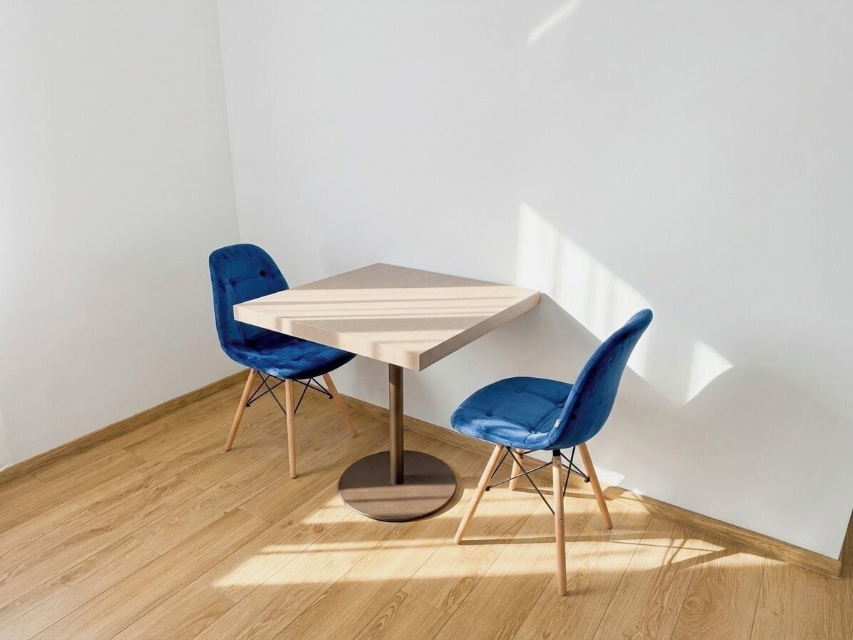 A room with a wooden floor, a light-colored table, and two blue chairs.