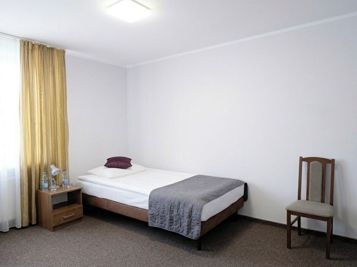 Een eenpersoonsbed met een witte matras, een grijze deken, een houten nachtkastje met een lamp en waterflessen, een houten stoel en een raam met gele gordijnen.
