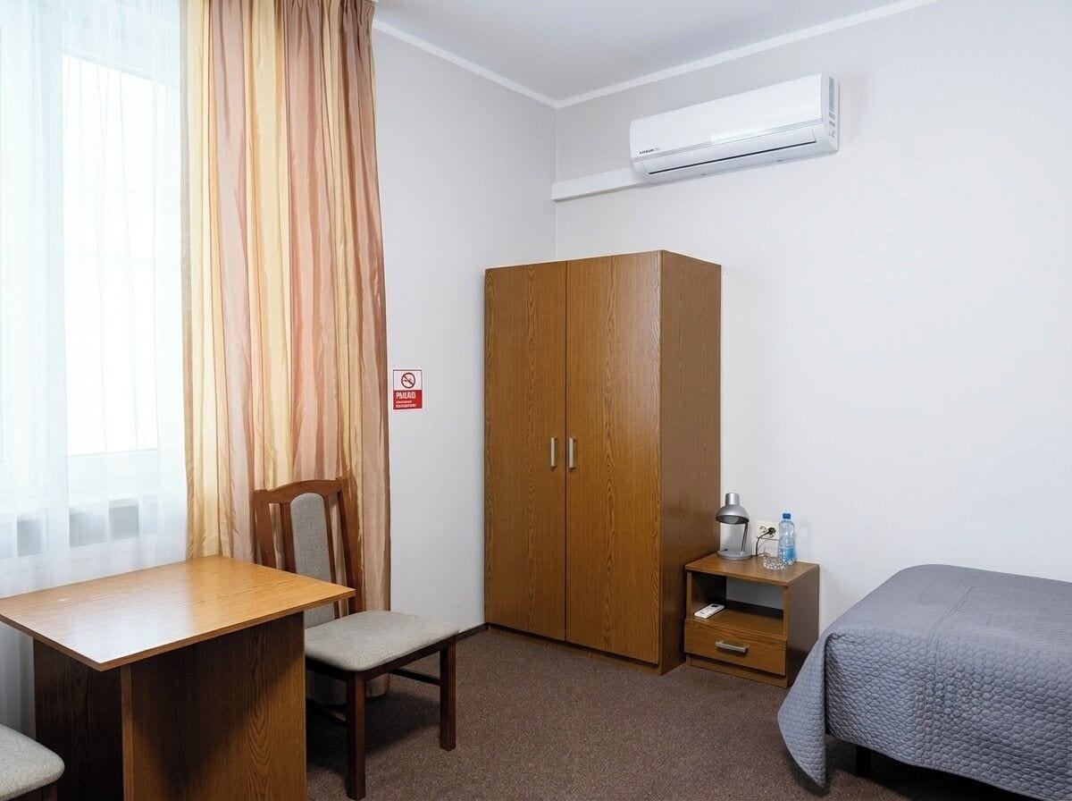 A hotel room with a wooden desk, a chair, a wardrobe, a bed, and an air conditioning unit.