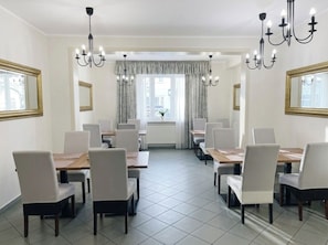 A dining area with multiple tables and chairs, chandeliers, and large windows with curtains.