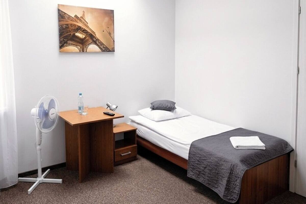 Ein kleines, ordentlich eingerichtetes Schlafzimmer mit einem Einzelbett, einem Holztisch, einem Standventilator, einer Wasserflasche und einem Bild des Eiffelturms.