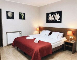A hotel room with a bed, two bedside lamps, a radiator, and three wall-mounted flower paintings.