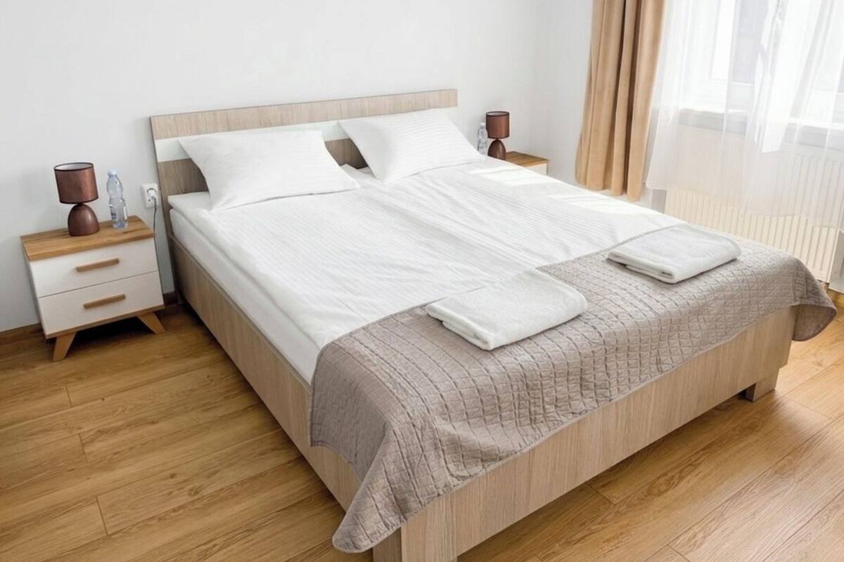Ein ordentlich bezogenes Bett mit einem Holzkopfteil, zwei Nachttische mit Lampen und ein Fenster mit transparenten Vorhängen.