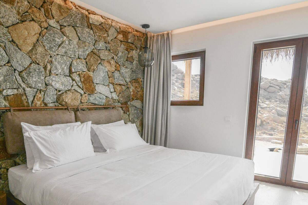 Quarto com parede de pedra, cama com roupa branca, janela e vista para o exterior.