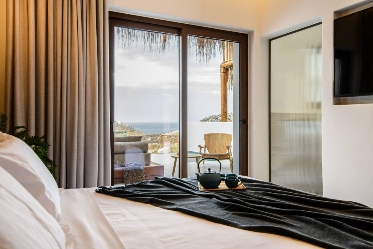 Quarto de hotel com uma cama, vista para o mar através de uma porta de correr de vidro, uma cadeira de madeira e uma pequena mesa com um bule de chá.