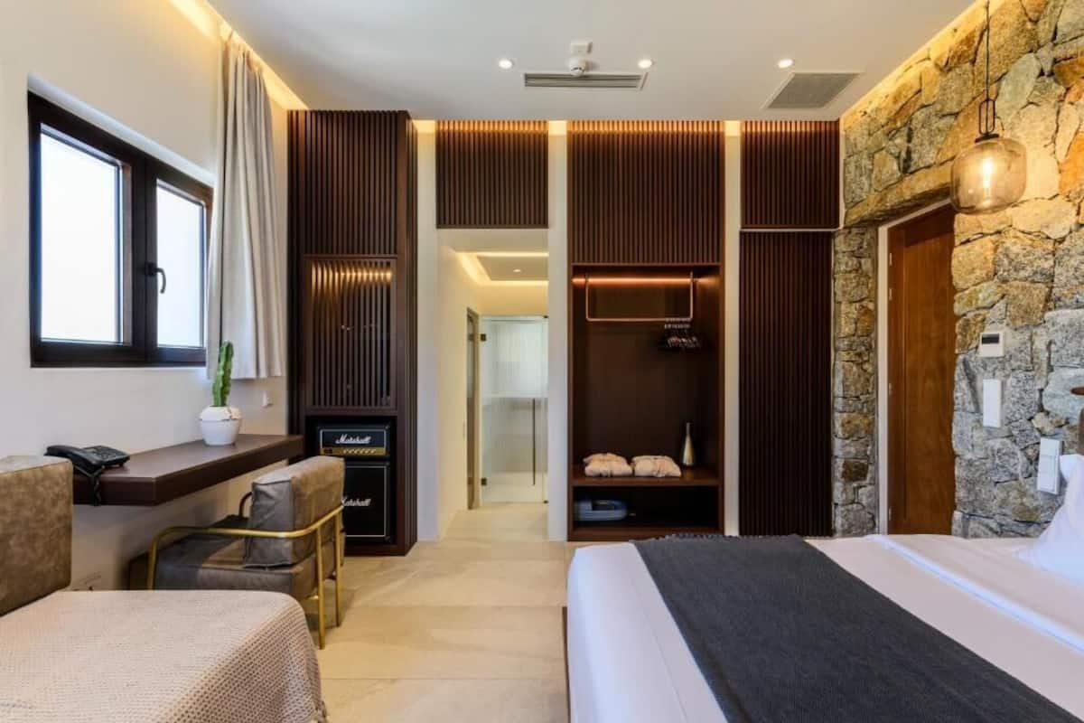 Quarto de hotel moderno com uma cama grande, área de estar com uma mesa pequena e cadeiras, sistema de som integrado e uma parede de detalhes em pedra.