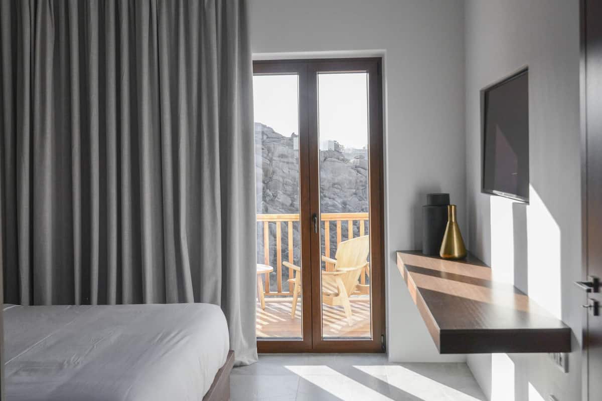 Quarto moderno com cama, porta de vidro deslizante para uma varanda, mesinha com um vaso e TV na parede.