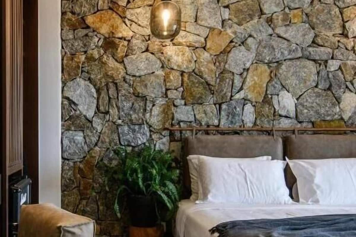 Um quarto com parede de pedra, uma cama com roupa de cama branca, um cobertor escuro, uma planta em vaso e um lustre.