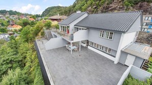 Une maison moderne à deux étages, dotée d’un patio couvert, d’un escalier menant à l’étage supérieur et offrant une vue sur une colline où se trouvent d’autres maisons.
