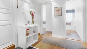 Un couloir aux murs blancs, une console blanche ornée d’objets décoratifs, un tapis gris et un tableau encadré accroché au mur.