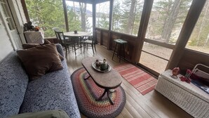 A cozy room with a blue patterned sofa, a round coffee table with a decorative item, and a view of trees through large windows. - Peaceful lake cottage in rolling hills of Maine (Sumner)