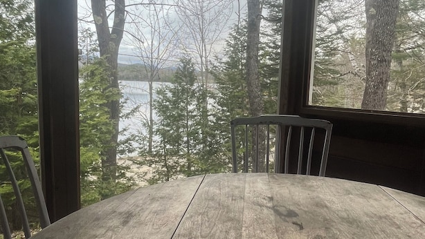 A wooden table in a covered outdoor area with a view of trees and a body of water. - Peaceful lake cottage in rolling hills of Maine (Sumner)