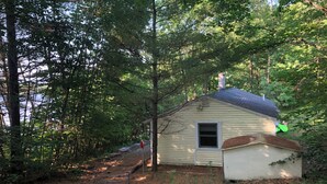 A small, single-story cabin with a sloped roof, surrounded by dense forest. A wooden staircase leads up to the entrance. - Peaceful lake cottage in rolling hills of Maine (Sumner)