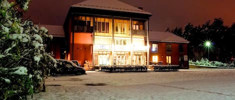 A two-story building with a snow-covered ground, a car parked in front, and a lit entrance.