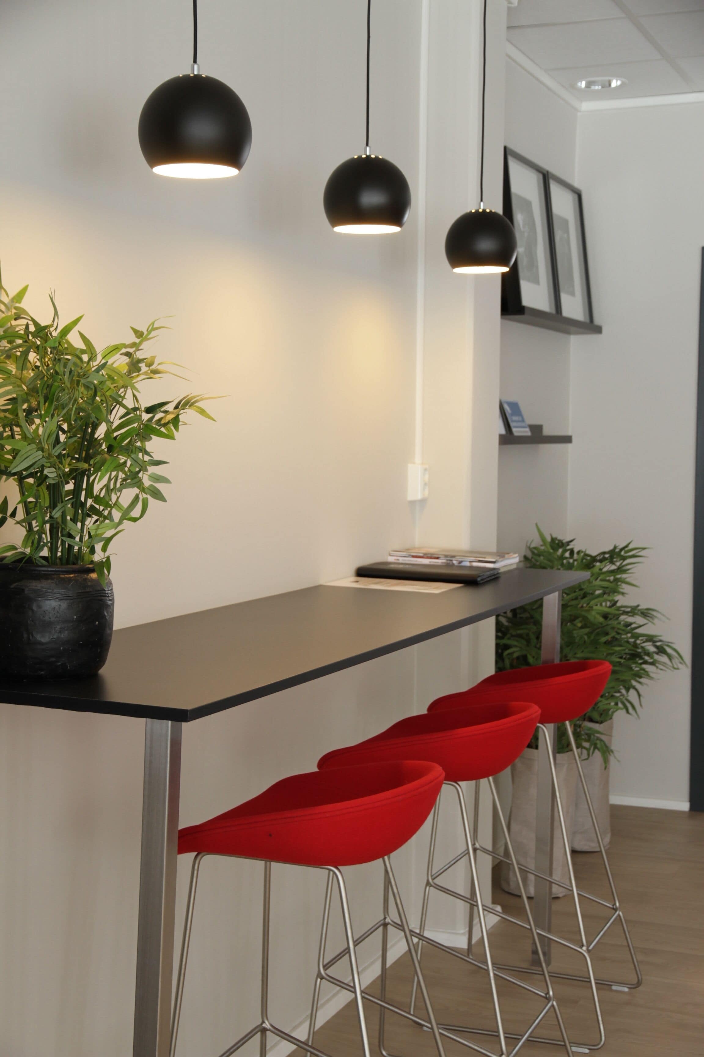 Een moderne keuken met een zwart aanrecht, drie rode barkrukken en hangende hanglampen.
