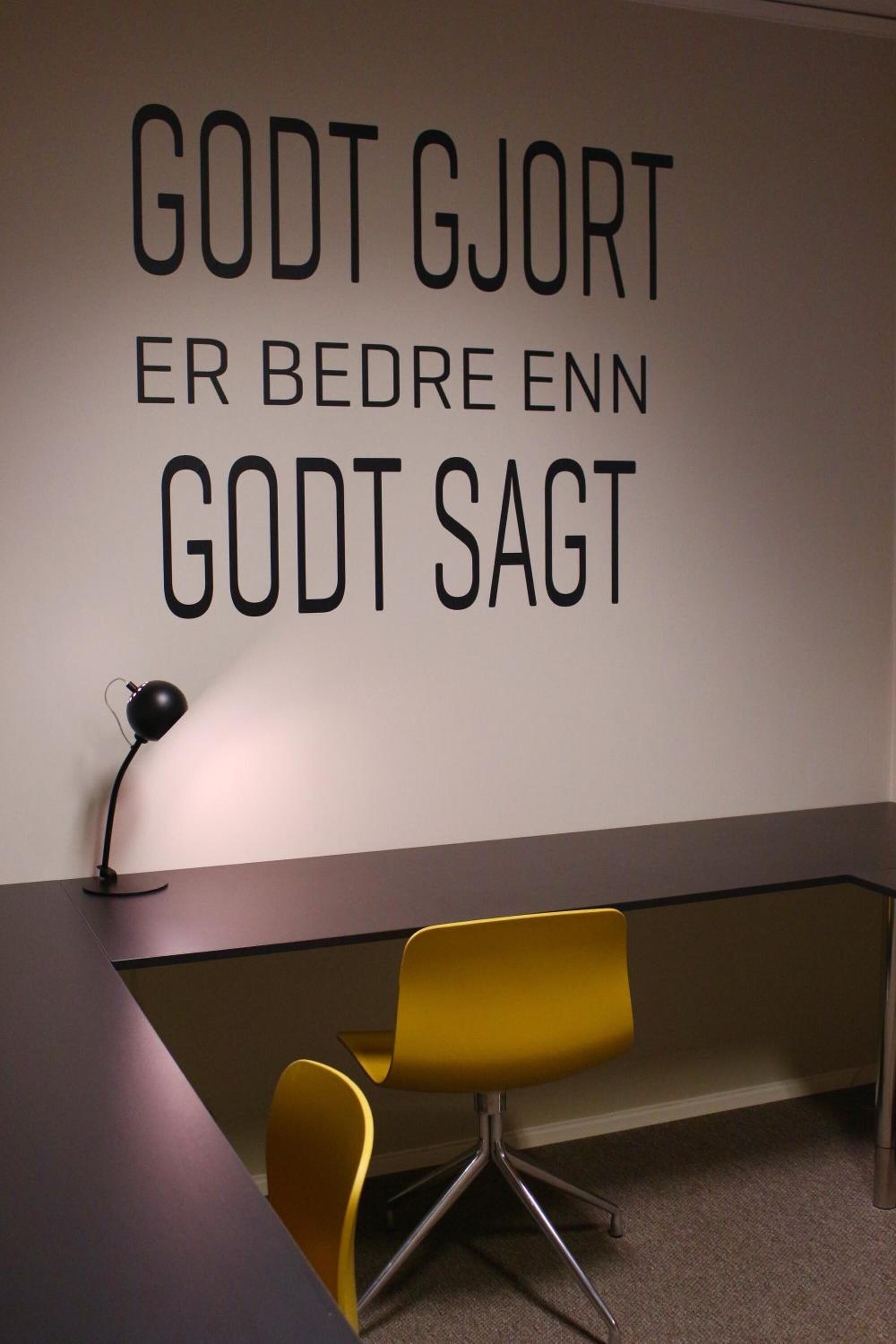Un espacio de oficina moderno con un escritorio, una silla amarilla y una pared con texto motivacional en un idioma extranjero.