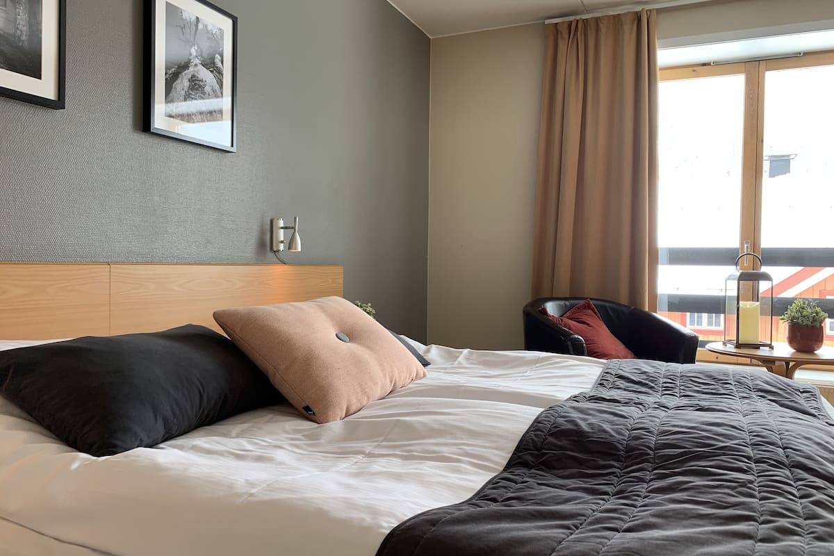 Habitación de hotel con cama, cabecero, lámpara de pared, una silla, ventana con cortinas y una mesita con planta.