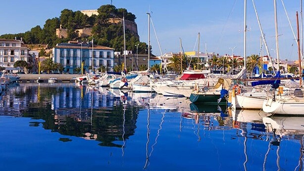 Un puerto deportivo con barcos amarrados, un edificio y una colina al fondo, bajo cielos despejados.