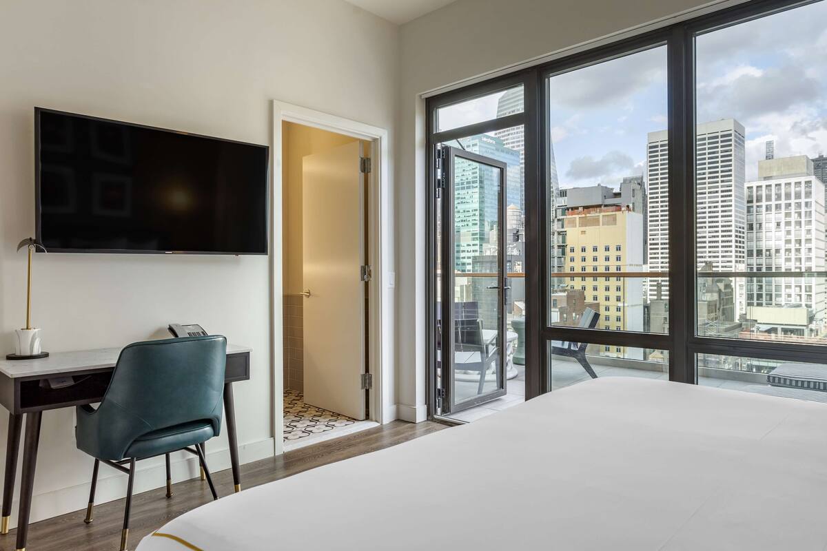 A hotel room with a bed, a desk with a chair, a TV, and a large window with a city view.
