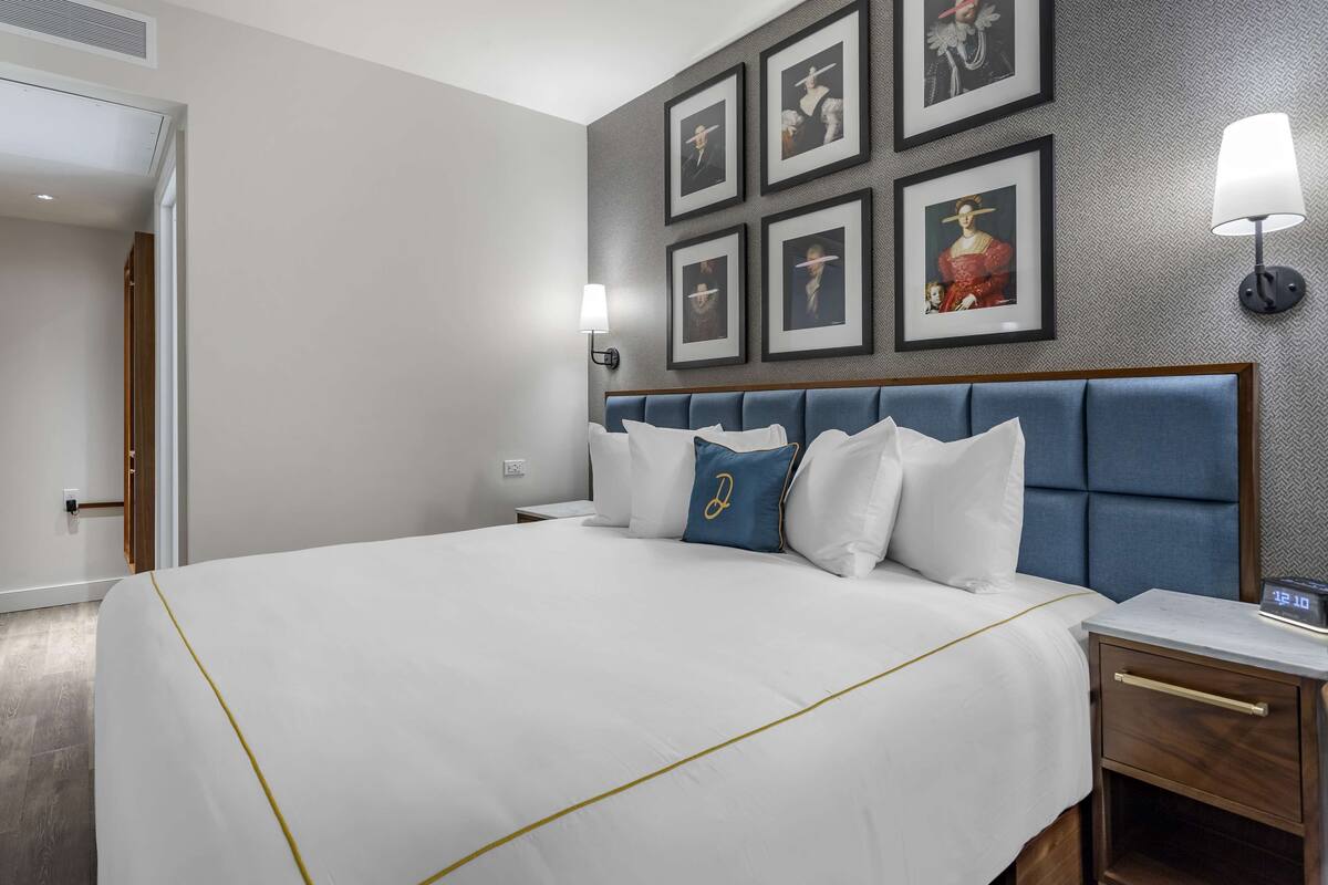 A hotel room with a large bed, a nightstand, a wall-mounted lamp, and framed artwork on the wall.