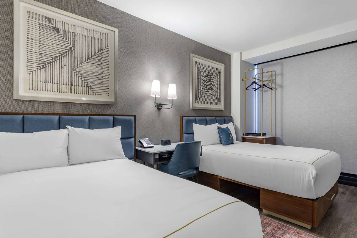 A hotel room with two beds, a desk, and a chair. There are wall-mounted lights and a coat rack.