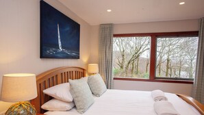 Habitación de hotel con una cama, dos almohadas, una mesita de noche con una lámpara, un cuadro de un velero y una ventana con vistas a árboles.