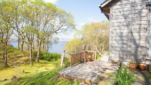 Una terraza de madera con vista a un lago, rodeada de árboles y una casa con porche.