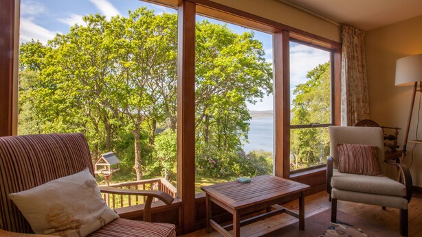 Habitación con amplias ventanas que dan a un lago y árboles, amueblada con sillas a rayas y una mesa de madera.