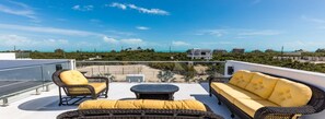 Eine Dachterrasse mit Korbmöbeln, einem runden Tisch und Blick auf Strand und Gebäude.