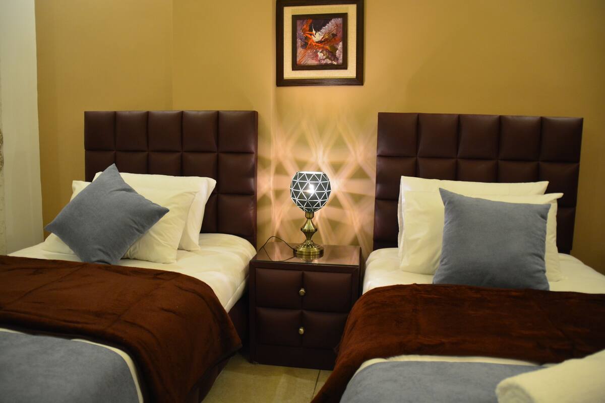 A hotel room with two beds, each with a brown headboard, a nightstand with a lamp, and a framed picture on the wall.