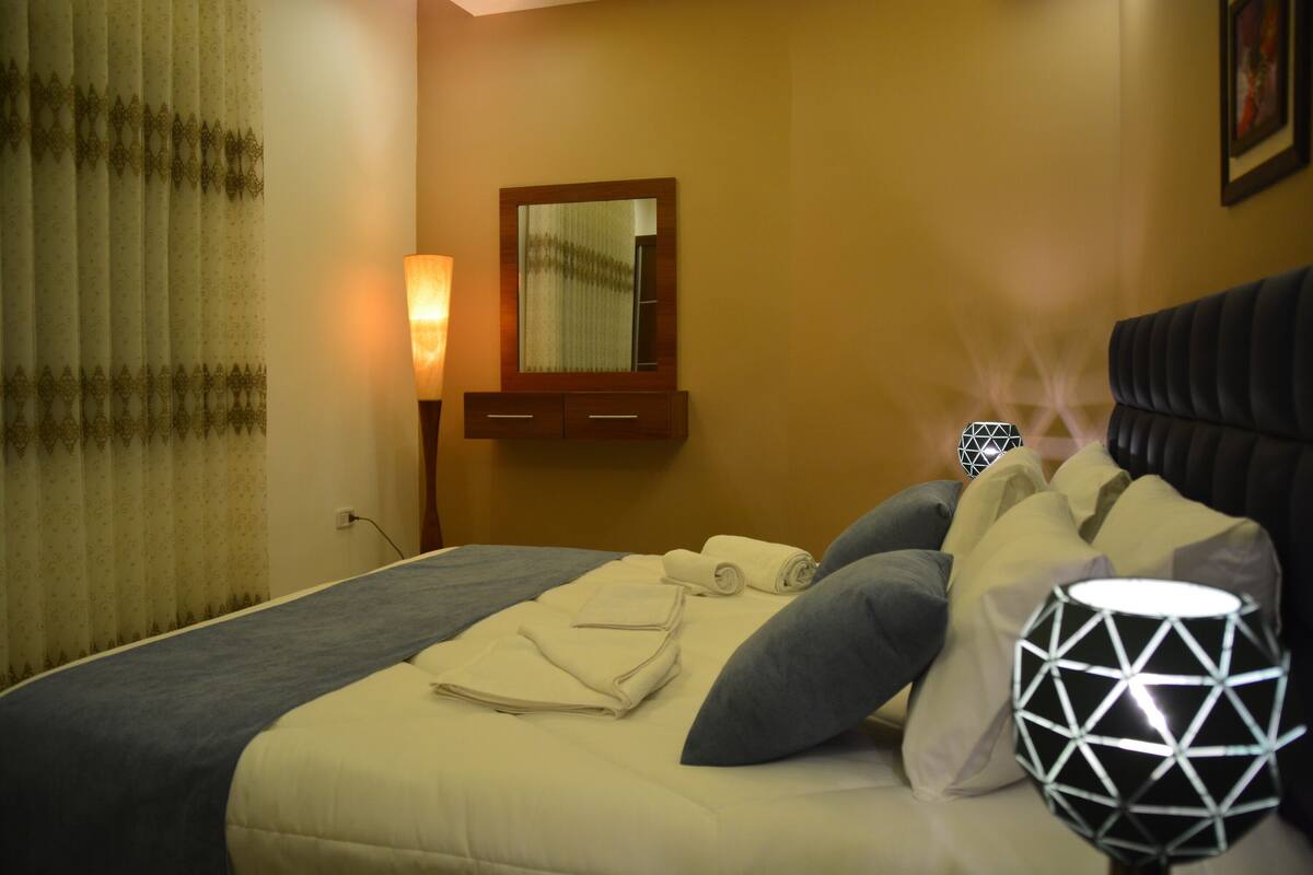 A neatly made bed with pillows and a rolled towel, a bedside lamp, and a geometric-patterned lamp on the nightstand.