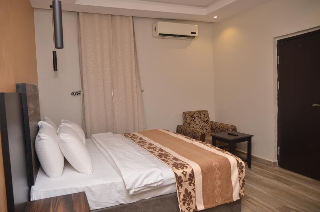 Quarto de hotel com uma cama, uma cadeira, uma mesinha, ar condicionado e uma porta.